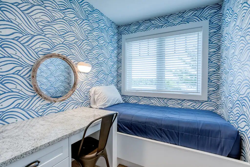 A single bed built into a window nook with blue wave-patterned wallpaper, a round mirror, and a built-in desk with a stool.