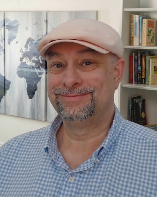 Robert Earl, wearing a flat cap, in front of a world map and bookshelves.